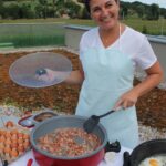 Ženska v beli majici in predpasniku stoji za mizo na prostem ter z lopatko meša slanino v električni ponvi. Ob njej so jajca in druga živila.