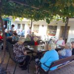 Skupina stanovalcev doma za starejše sedi pod pergolo na dvorišču in zajtrkuje ob lesenih mizah, obdani z zelenjem vinske trte.