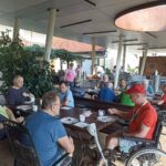 Moški stanovalci doma za starejše zajtrkujejo na pokriti terasi ob dolgih lesenih mizah; več jih sedi v invalidskih vozičkih.