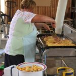 Zaposlena pripravlja umešana jajca na večji kuhinjski plošči na prostem, spredaj so zloženi krožniki in porcije pripravljene hrane.