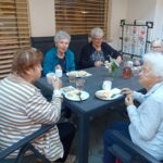 Štiri starejše ženske sedijo za mizo v atriju doma in zajtrkuje; na mizi so krožniki, skodelice in vrči z napitki.