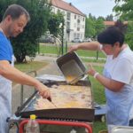 Moški meša hrano na veliki zunanji plošči, ženska poleg njega vliva tekočino v ponev med pripravo zajtrka na prostem.
