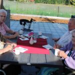 Štiri starejše osebe sedijo za zunanjo mizo, obloženo z rdečim pogrinjkom in skodelicami. Sproščeno se pogovarjajo in pijejo kavo ob cvetličnem aranžmaju.