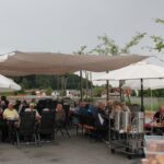 Skupina starejših oseb sedi za mizami na zunanji terasi doma starejših, pod senčniki in platnenimi nadstreški, ter zajtrkuje v prijetnem zunanjem okolju.
