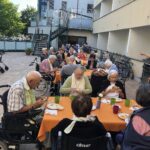 Starejši stanovalci doma za starejše sedijo na terasi za dolgimi mizami z oranžnimi prti in zajtrkujejo.