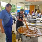 Dva moška pripravljata golaž v veliki posodi na prostem, v ozadju sedijo starejši ljudje in opazujejo dogajanje.