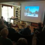 Skupina starejših oseb sedi v poltemnem prostoru in gleda projekcijo filma na velikem platnu; na zaslonu je prizor motoristov in ljudi pred gradom.