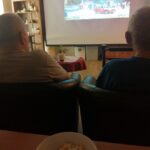 Pogled izza dveh starejših moških, ki sedita v naslanjačih in gledata film; v ospredju je skleda s pokovko, na platnu pa prizor z avtomobili in ljudmi pred gradom.