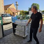 Ženska in moški na dvorišču potiskata voziček, naložen s plastenkami vode, v ozadju je vhod v stavbo doma.