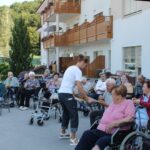 Zaposlena deli pečeno koruzo stanovalcem doma, ki sedijo zunaj na dvorišču, nekateri v invalidskih vozičkih, drugi na stolih.