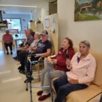 Skupina stanovalcev doma za starejše sedi v vrsti ob steni skupnega prostora, nekateri uporabljajo hodulje ali invalidske vozičke. V ospredju ženska v rožnatem puloverju drži glasbilo – ropotuljo.