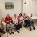 Stanovalci doma za starejše sedijo ob steni in mizah v skupnem prostoru. V ospredju tri ženske sedijo skupaj, ena od njih drži hoduljo z rumeno rožo.