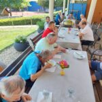 Moški stanovalci doma za starejše kosijo zunaj za dolgo mizo; jedo juho, na mizi so kozarci in cvetlični aranžma.