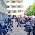 Skupina stanovalcev doma za starejše sedi na dvorišču med telovadbo; dvignjene roke proti nebu, ob strani so hodulje in invalidski vozički.