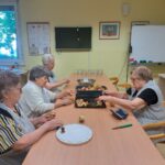 Štiri starejše gospe sedijo ob dolgi mizi in čistijo ter režejo jabolka. Na mizi so krožniki, noži in sklede s sadjem.