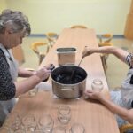Dve starejši gospe z zajemalko polnita kozarce z vročo marmelado iz aronije iz velike kovinske posode. Na mizi je več praznih steklenih kozarcev.
