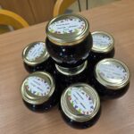 Sedem kozarcev domače marmelade iz aronije, z zlatimi pokrovčki in pisanimi etiketami, zloženih v piramido na leseni mizi.