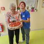 Stanovalka s posodo paradižnikov v rokah, ob njej zaposlena v modri majici, ki se smehlja proti kameri.