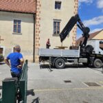 Moški nadzoruje dvigovanje velike lesene kolutne mize s tovornjaka z avtodvigalom na parkirišču pred staro stavbo z okrašenim robom.