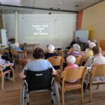 Projekcija filma v dvorani doma upokojencev, stanovalci so obrnjeni proti platnu, v ospredju je oseba na invalidskem vozičku.