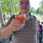 Moški v črtasti majici stoji na terasi in dviguje kozarec z osvežilnim sadnim napitkom.