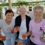 Tri nasmejane starejše gospe držijo v rokah kozarce z osvežilnim sadnim napitkom. Stojijo na terasi doma z vinogradom v ozadju.