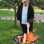Starejši moški na vrtu dodaja poleno na ogenj v kovinskem kurišču, okoli njega je zelenica in drevo s plodovi.