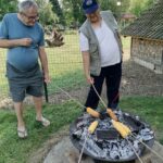 Dva moška pečeta koruzo nad ognjem v kovinskem kurišču, eden drži palico, drugi nadzira pečenje.