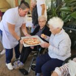 Oskrbovanka v invalidskem vozičku prejema kos pice od zaposlene, ki na pladnju drži sveže pečeno pico, ob njej sedi še ena oskrbovanka.