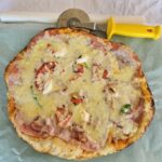 Sveže pečena pica z raztopljenim sirom, šunko in zelenjavo na peki papirju, ob njej rezalnik za pico z rumenim ročajem.
