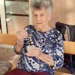 Starejša gospa s kratkimi sivimi lasmi, oblečena v modro-belo vzorčasto majico, sedi zunaj na leseni klopi in z žlico zajema ledeno kavo iz kozarca.