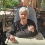 Starejša gospa s kratkimi belimi lasmi, oblečena v črno majico, sedi v invalidskem vozičku za mizo na terasi in drži kozarec ledene kave, medtem ko jo obsije sonce.