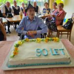 Moški v vozičku sedi za torto z napisom »90 LET«, okoli njega so gostje.