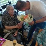 Starejša gospa v vozičku sprejema šopek cvetja od moškega, ki se je sklonil k njej in ji stisnil roko. Na mizi pred njima so kozarec pijače, prtiček in vilice. Gospa se nasmejano ozira proti moškemu.