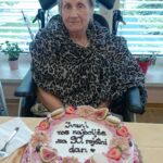 Starejša gospa v vozičku sedi za mizo in gleda v kamero. Pred njo je rojstnodnevna torta, okrašena s sadjem, makroni in rožami. Na torti piše: »Ivani vse najboljše za 90. rojstni dan.«