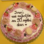 Rojstnodnevna torta na rumeni podlagi. Okrašena je z rožnatimi smetanastimi obrobami, figami, jagodami, borovnicami, makroni in sladkornimi rožami. Na sredini je napis: »Ivani vse najboljše za 90. rojstni dan.«