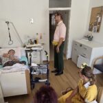 Klovnovski par stoji ob postelji stanovalke v sobi, eden stoji, drugi sedi na stolu, stanovalka pa leži v postelji in jih posluša.
