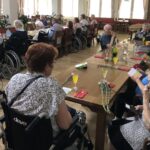 Skupina stanovalcev doma za starejše sedi za dolgo okrašeno mizo, na kateri so napitki in rože, ter skupaj praznuje rojstne dni.