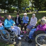 Skupina starejših žensk, od katerih sta dve v invalidskem vozičku, sedi v krogu na klopi in se druži v senci dreves v domu za starejše.