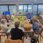 Skupina starejših oseb in mentorica sedijo za mizo ter izdelujejo sončnice iz papirja. Na mizi so šopki rumenih cvetov in košara s slamo.