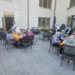 Stanovalci doma upokojencev zajtrkujejo zunaj na terasi, sede za mizami z oranžnimi prti, osebje pa postreže hrano.