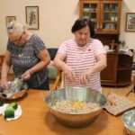 Dve gospe pripravljata maso iz naribanih bučk in jajc v velikih kovinskih skledah, tretja sedi ob strani.