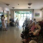 Ženska v beli obleki stoji pred skupino stanovalcev doma upokojencev in jim govori v mikrofon.