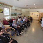 Stanovalci doma upokojencev, večinoma na invalidskih vozičkih, poslušajo žensko v beli obleki, ki jim govori v mikrofon.