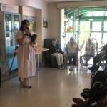 Ženska v beli obleki govori v mikrofon in drži punčko, medtem ko skupina stanovalcev sedi in posluša.