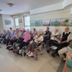 Večja skupina stanovalcev doma upokojencev sedi v vrsti in pije čaj, medtem ko poslušajo nastop.