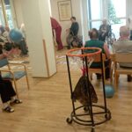 Skupina starejših oseb sedi v krogu na stolih in vozičkih. St starejša ženska v ospredju meče modro žogo proti premični košari na stojalu. Ostali gledajo in čakajo na vrsto.