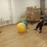 Tri starejše ženske sede na stolih v prostoru z lesenim podom. Ena ima v rokah modro žogo, druga gleda proti velikim žogam na tleh (modra in rumena). V ozadju so škatle in fitnes naprave.