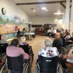 Skupina stanovalcev doma za starejše sedi v dvorani in posluša literarni dogodek. Na steni je poslikava mesta Ptuj, v ospredju pa dve govornici za mizo.