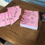 Knjige z rožnatimi platnicami, z naslovom Tako to počnejo volkovi avtorice Kaje Teržan, razstavljene na leseni mizi. Ob strani so programske knjižice festivala Dnevi poezije in vina.